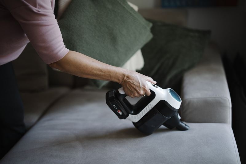 A person is holding a handheld vacuum cleaner while cleaning a couch. The scene takes place in a living room with green cushions. Sunlight is coming through a w